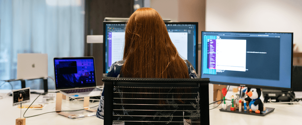 a person with long hair works at a desk with two monitors and a laptop